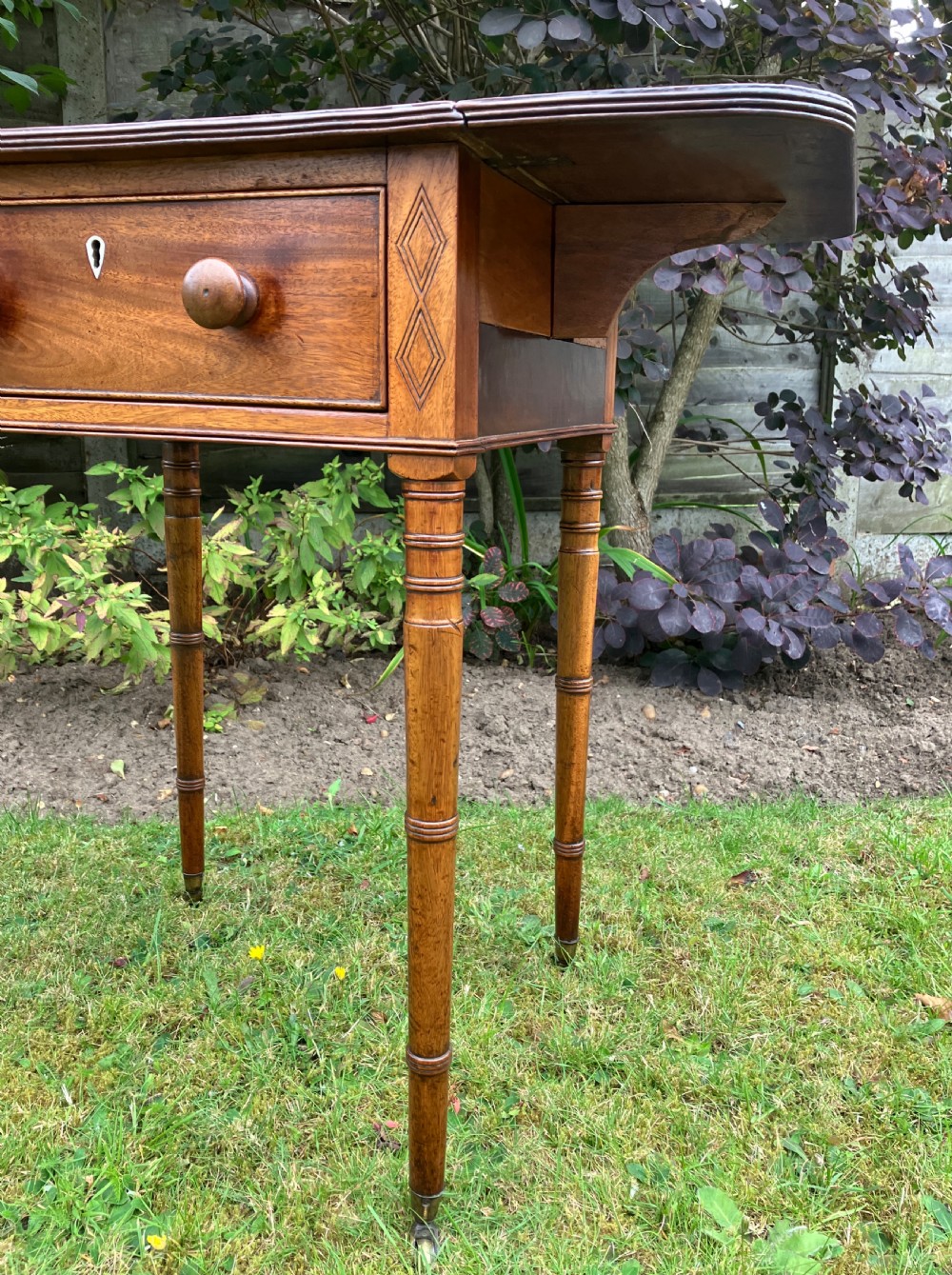 A late Georgian mahogany work table featuring twin drop leaves, a single drawer with wooden knobs, inlaid diamond details, and turned legs on brass castors. View VII