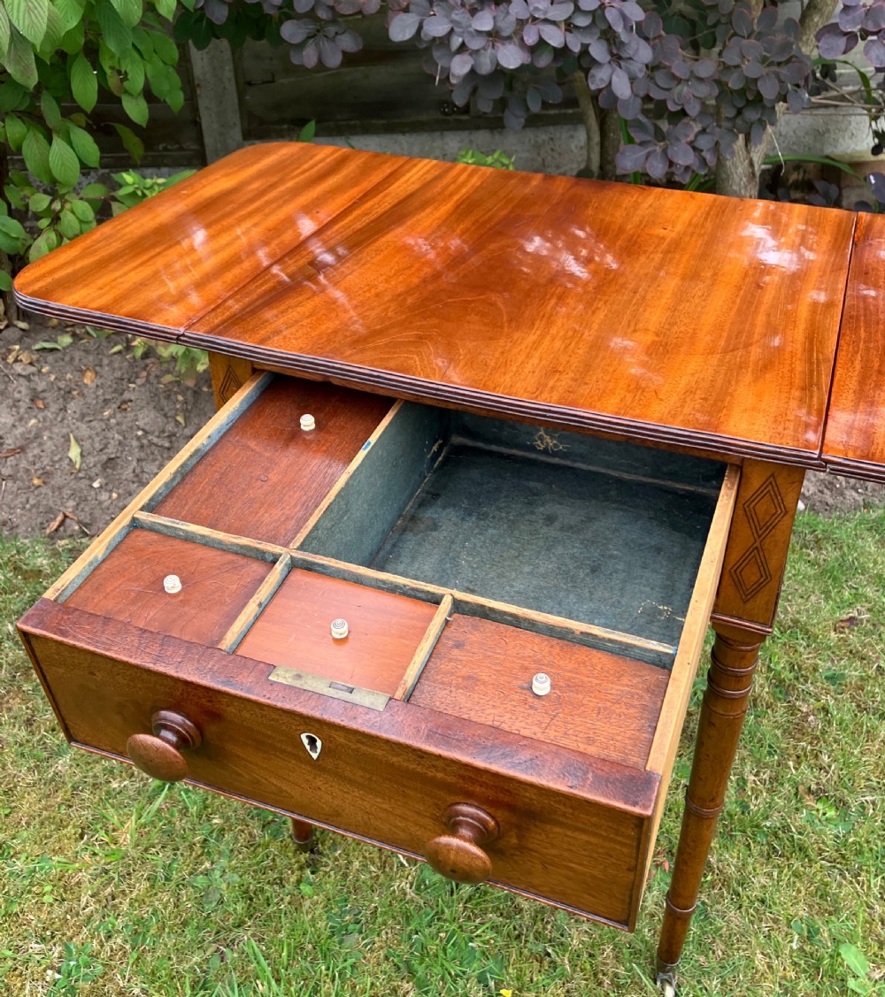 A late Georgian mahogany work table featuring twin drop leaves, a single drawer with wooden knobs, inlaid diamond details, and turned legs on brass castors. View IX