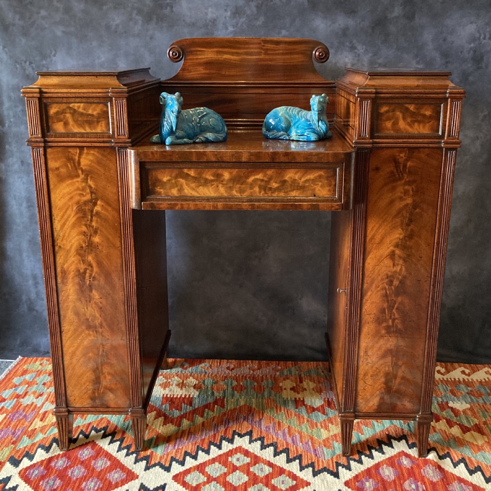 An Early Victorian flame mahogany pedestal sideboard with a central recessed drawer section, scrolled backboard, fluted pilasters, and twin side cupboard doors.
