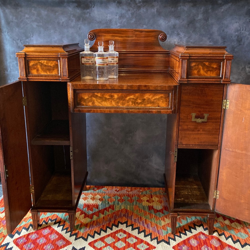 An Early Victorian flame mahogany pedestal sideboard with a central recessed drawer section, scrolled backboard, fluted pilasters, and twin side cupboard doors. View VI