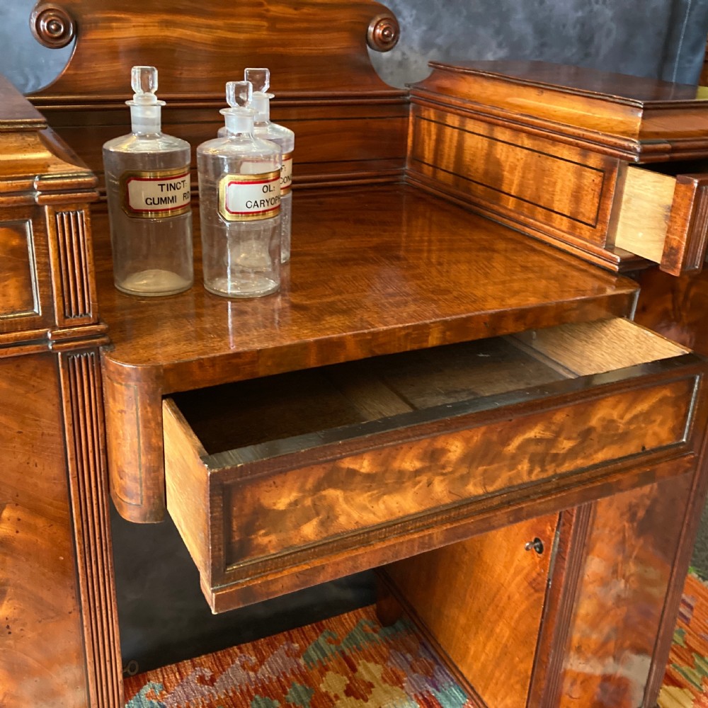 An Early Victorian flame mahogany pedestal sideboard with a central recessed drawer section, scrolled backboard, fluted pilasters, and twin side cupboard doors. View XV