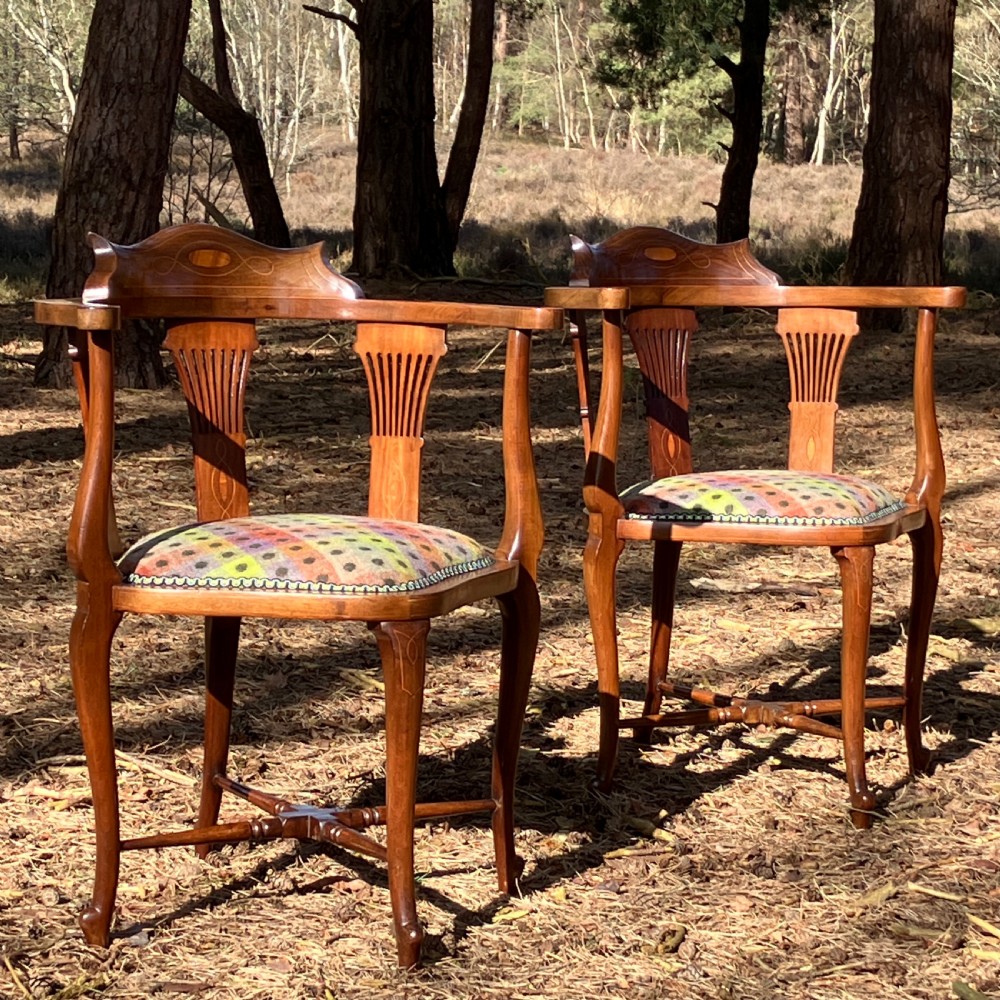A pair of Edwardian mahogany corner chairs featuring curved top rails with marquetry inlay, vertical splats, upholstered patterned seats, and slender legs joined by stretchers.