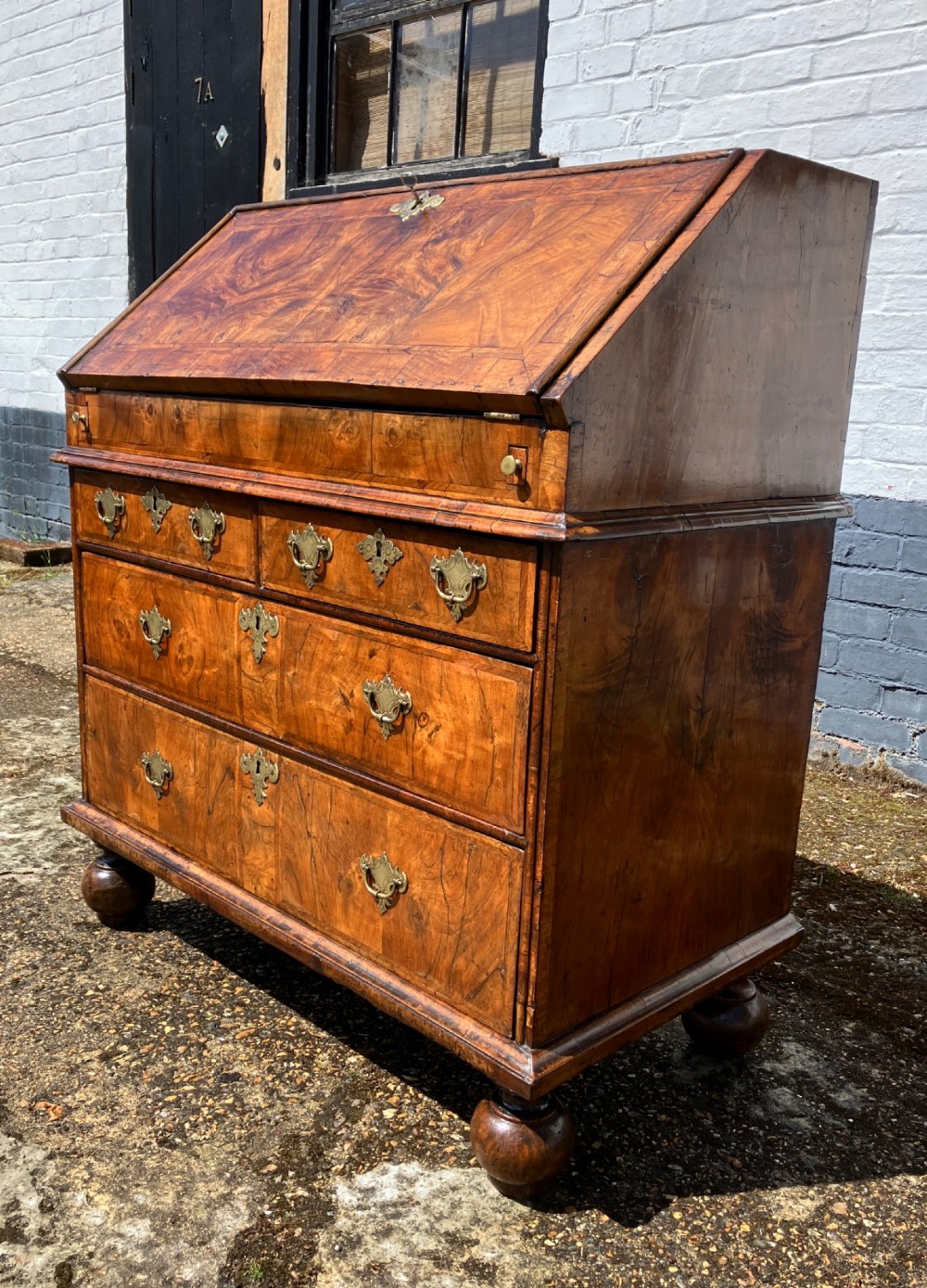 An early eighteenth-century English walnut fall-front bureau featuring burr veneer panels, decorative brass escutcheons and handles, and turned bun feet. View III