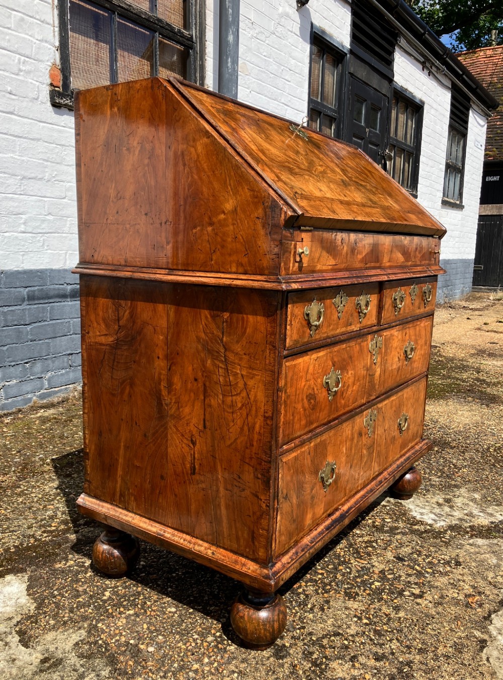 An early eighteenth-century English walnut fall-front bureau featuring burr veneer panels, decorative brass escutcheons and handles, and turned bun feet. View IV