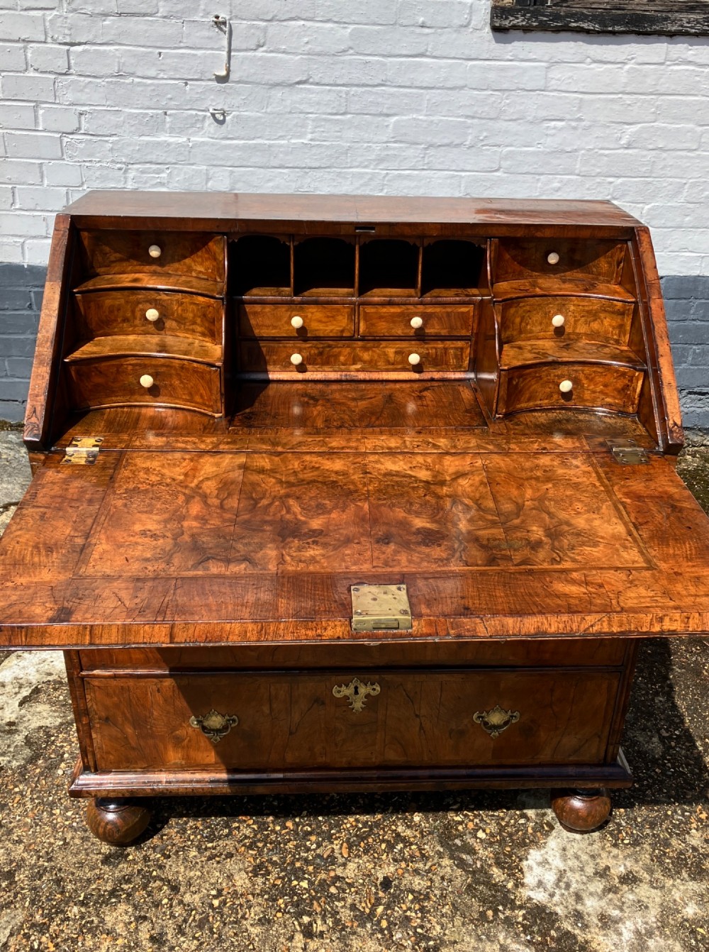 An early eighteenth-century English walnut fall-front bureau featuring burr veneer panels, decorative brass escutcheons and handles, and turned bun feet. View V