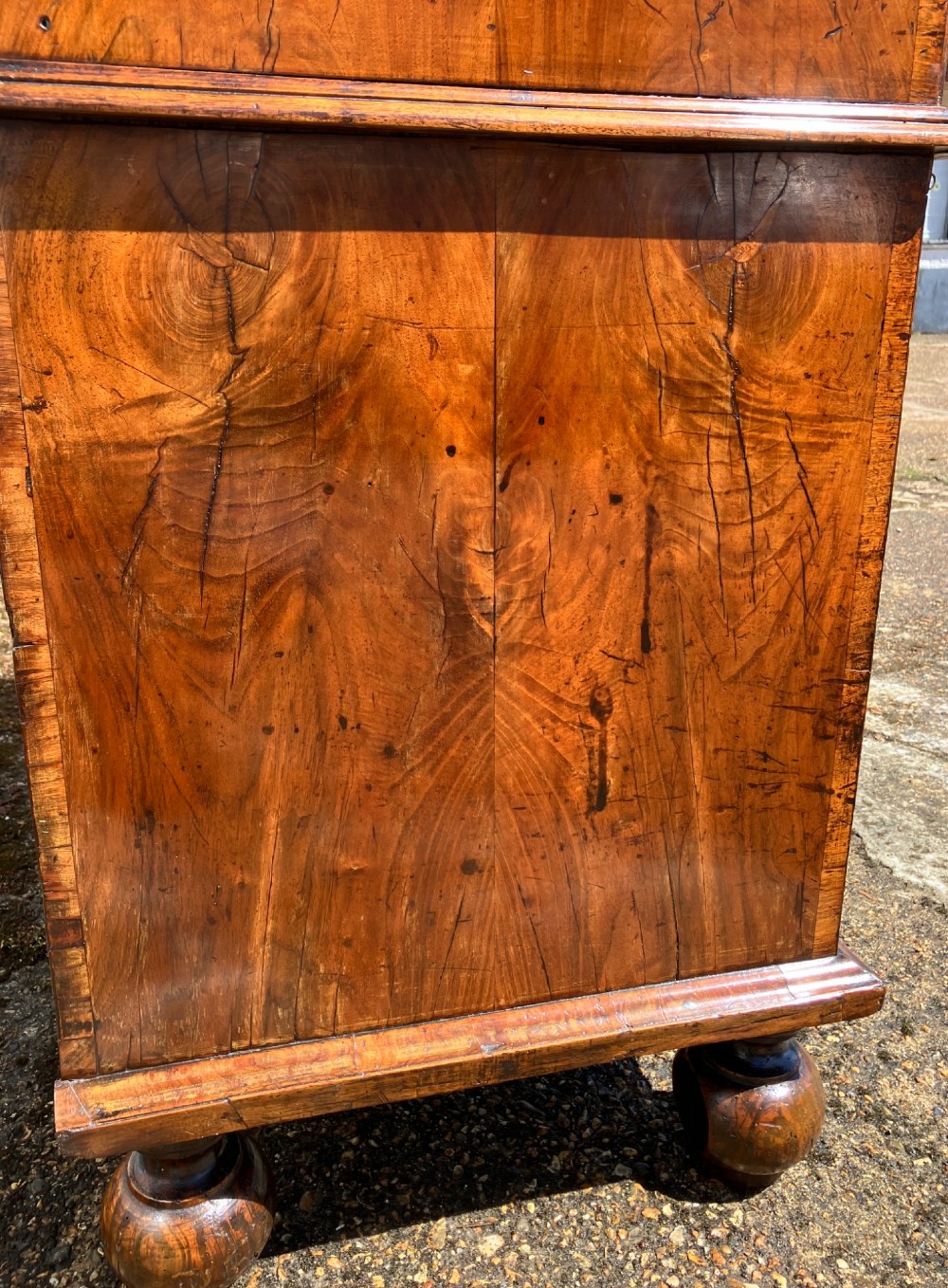 An early eighteenth-century English walnut fall-front bureau featuring burr veneer panels, decorative brass escutcheons and handles, and turned bun feet. View X