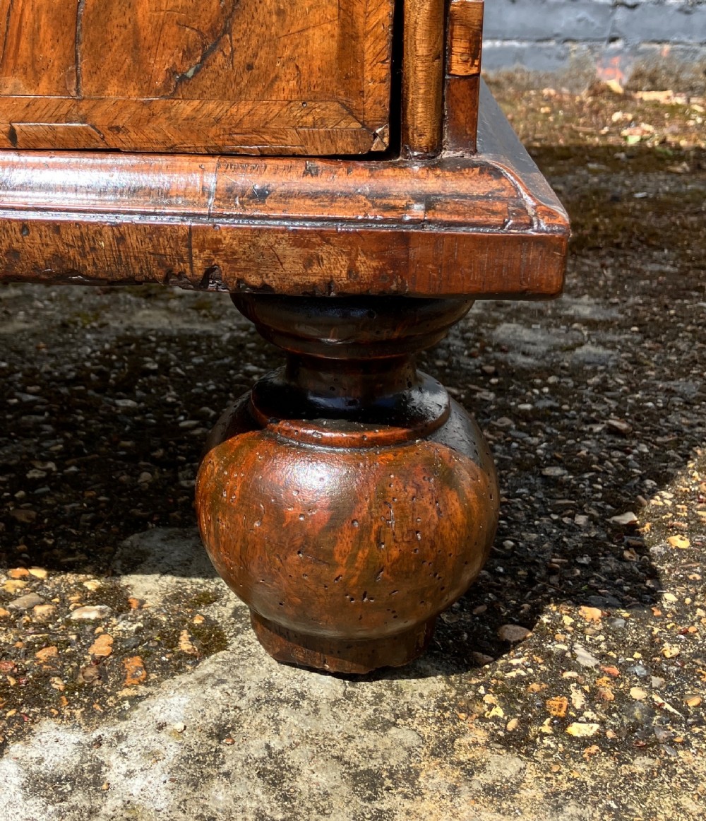 An early eighteenth-century English walnut fall-front bureau featuring burr veneer panels, decorative brass escutcheons and handles, and turned bun feet. View XIII
