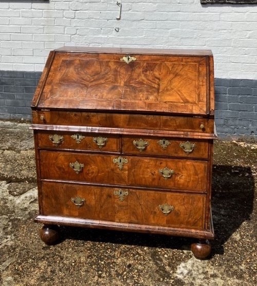 An early eighteenth-century English walnut fall-front bureau featuring burr veneer panels, decorative brass escutcheons and handles, and turned bun feet. View II