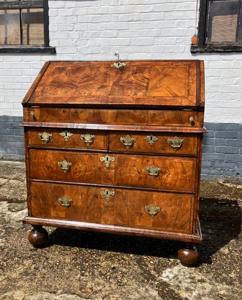 An early 18th-century English walnut fall front bureau featuring figured timber veneers, brass handles, and turned bun feet.