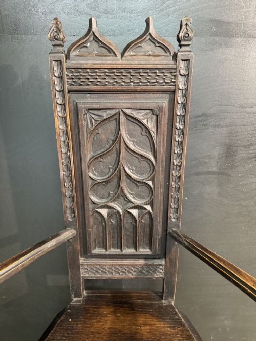 An antique dark oak open armchair featuring a tall carved back panel decorated with Gothic tracery motifs, curved armrests, and turned front legs. View II