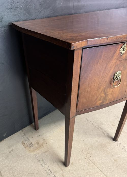 An antique Georgian mahogany bow-front sideboard featuring a central drawer, flanking cupboard doors, brass lion mask ring handles, and square tapered legs. View X