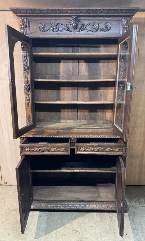 A two-tier dark oak cabinet featuring arched glazed top doors, carved scroll motifs, and lower doors with relief carvings of game birds. View VI