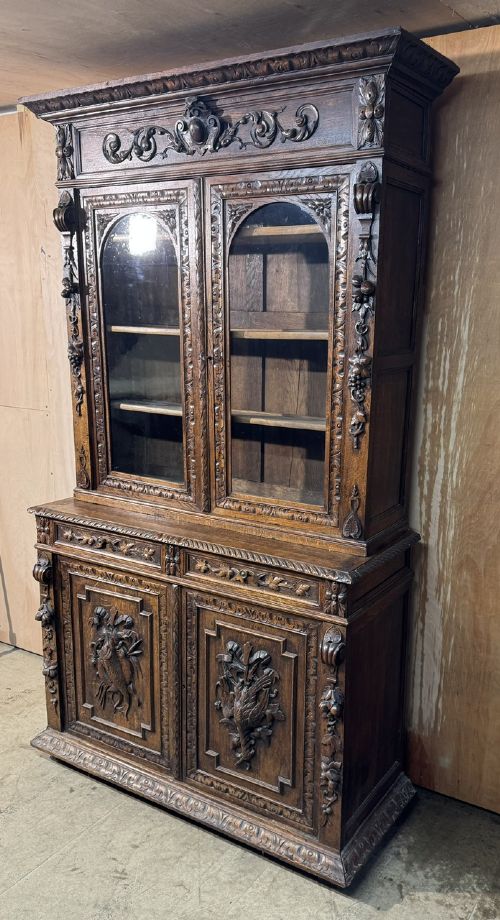 A two-tier dark oak cabinet featuring arched glazed top doors, carved scroll motifs, and lower doors with relief carvings of game birds. View X