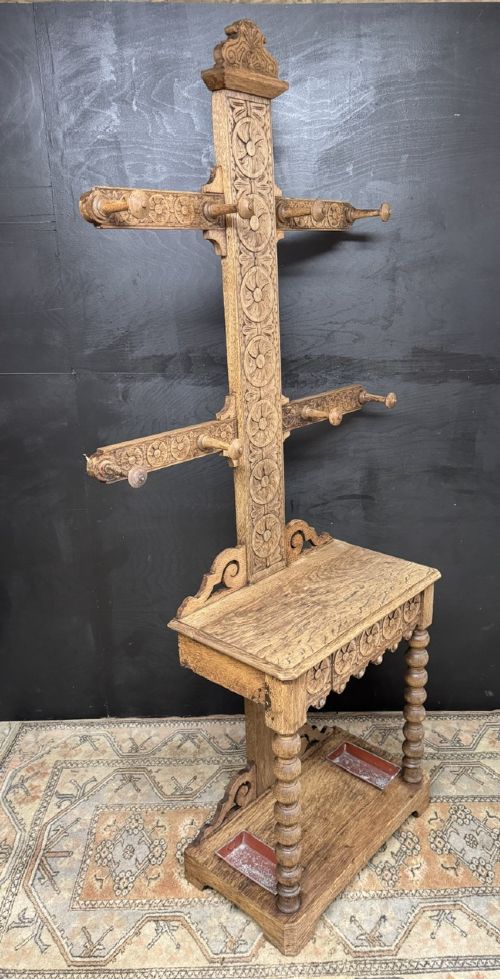 A tall carved oak hall stand featuring two horizontal rails of turned coat pegs, a central column carved with floral rosettes, bobbin-turned legs, and lower umbrella drip trays. View VII