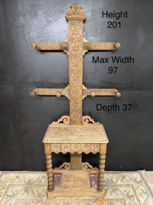A tall carved oak hall stand featuring two horizontal rails of turned coat pegs, a central column carved with floral rosettes, bobbin-turned legs, and lower umbrella drip trays. View XII
