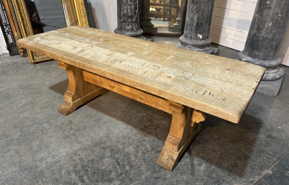 A rustic bleached oak dining table featuring a thick rectangular top supported by carved trestle ends connected with a stretcher beam. View XXIII