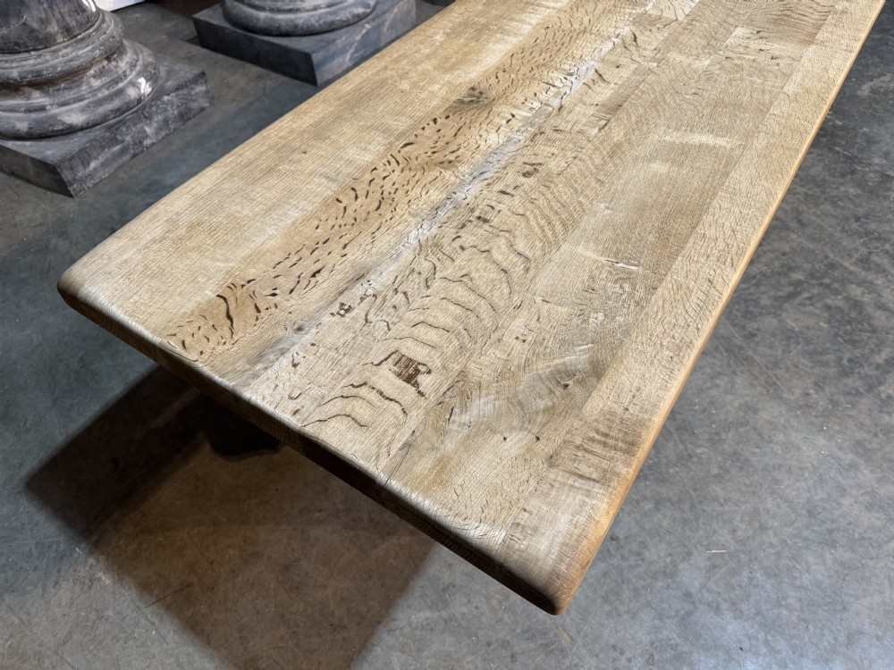 A rustic bleached oak dining table featuring a thick rectangular top supported by carved trestle ends connected with a stretcher beam. View XXV