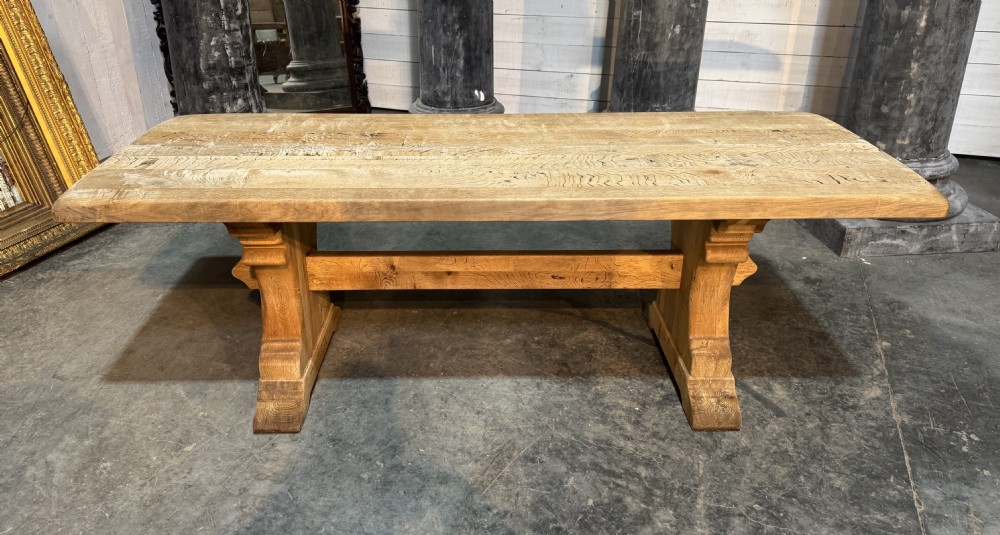 A rustic bleached oak dining table featuring a thick rectangular top supported by carved trestle ends connected with a stretcher beam. View XXVII