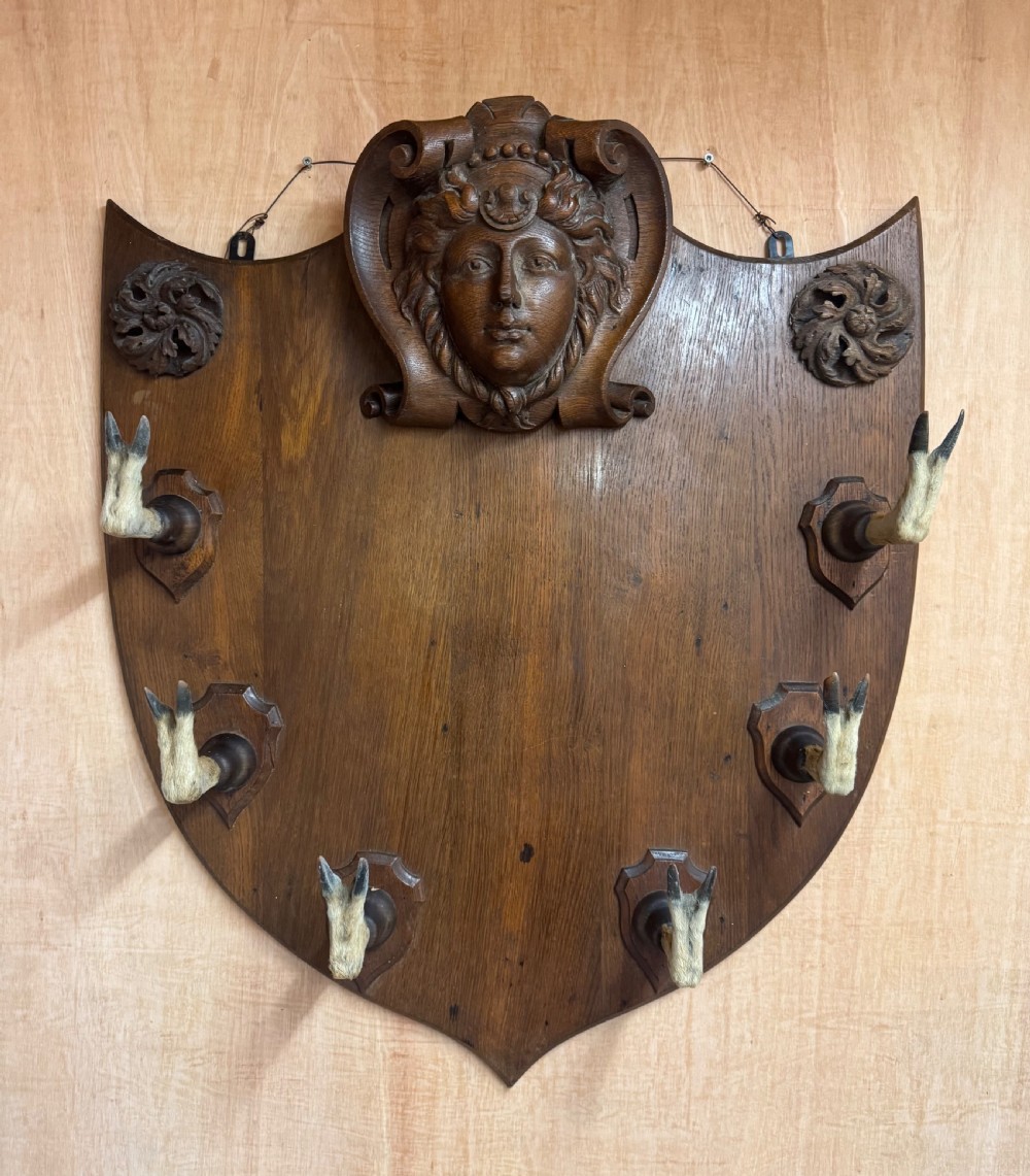 A shield-shaped carved oak wall rack featuring a central carved face, decorative rosettes, and six mounted deer hoof hooks.