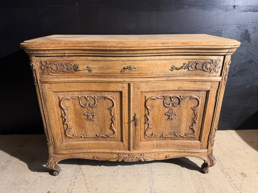 An antique French carved oak cabinet featuring a single top drawer above two panelled doors with carved scroll motifs and brass hardware, supported by short cabriole feet.