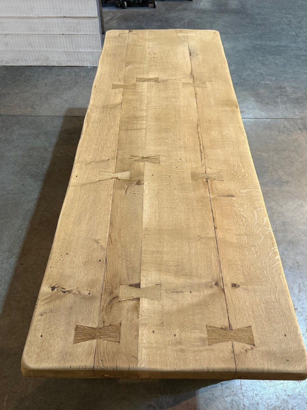 A large light oak farmhouse dining table featuring a solid plank top reinforced with butterfly joints and a sturdy trestle base connected by a central stretcher. View X