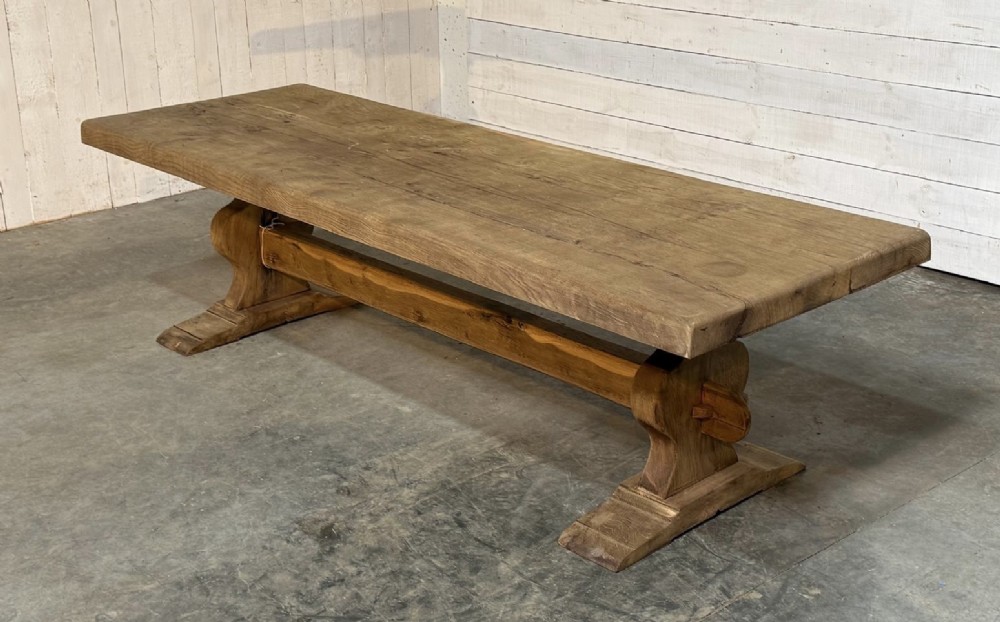 A large rustic French oak dining table featuring a thick timber plank top supported by a sturdy trestle base with a central keyed stretcher.