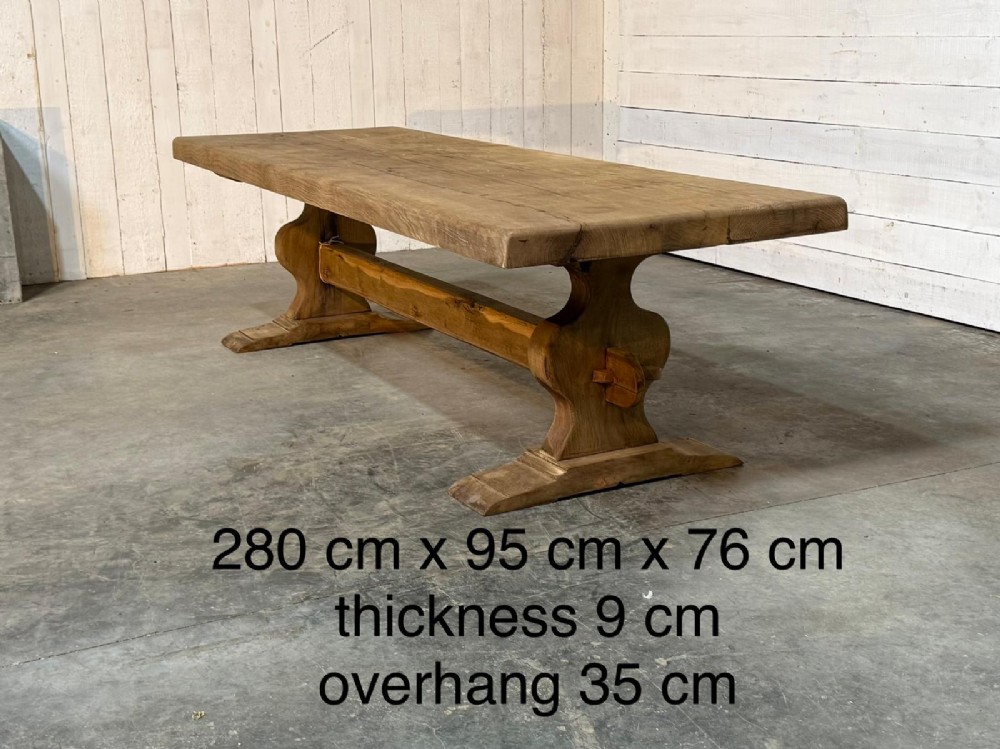 A large rustic French oak dining table featuring a thick timber plank top supported by a sturdy trestle base with a central keyed stretcher. View XII