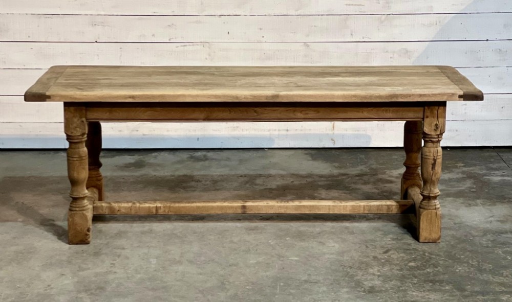 A rectangular bleached oak farmhouse dining table featuring turned legs, a low central stretcher, and a breadboard top.