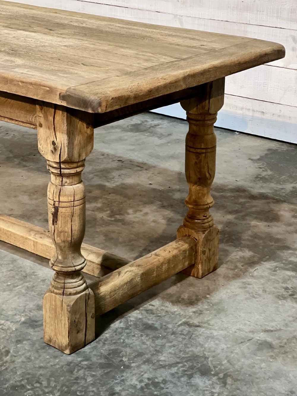 A rectangular bleached oak farmhouse dining table featuring turned legs, a low central stretcher, and a breadboard top. View VI