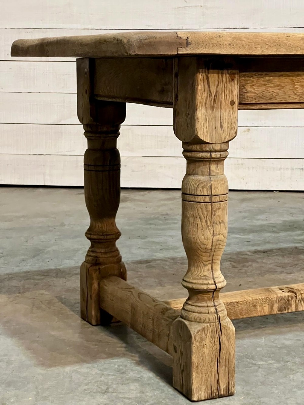 A rectangular bleached oak farmhouse dining table featuring turned legs, a low central stretcher, and a breadboard top. View XII