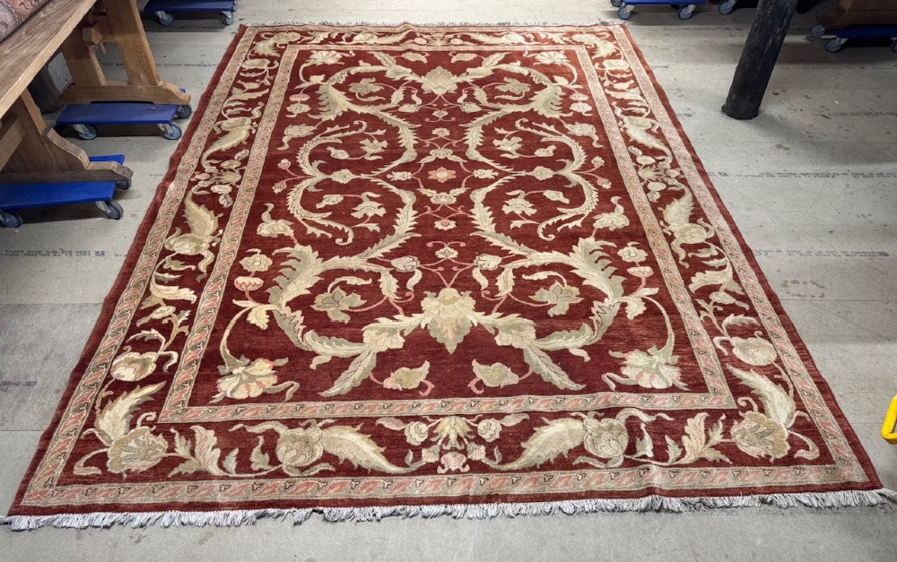 A large woven wool carpet featuring a deep red field decorated with cream and sage green floral scroll motifs and enclosed within a matching multi-tiered border.