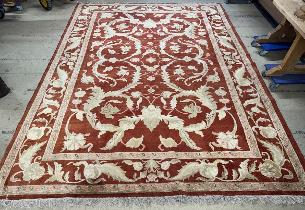 A large woven wool carpet featuring a deep red field decorated with cream and sage green floral scroll motifs and enclosed within a matching multi-tiered border. View IX