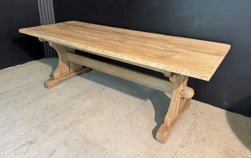A long bleached oak dining table featuring a solid rectangular top supported by curved trestle legs connected by a central stretcher beam.