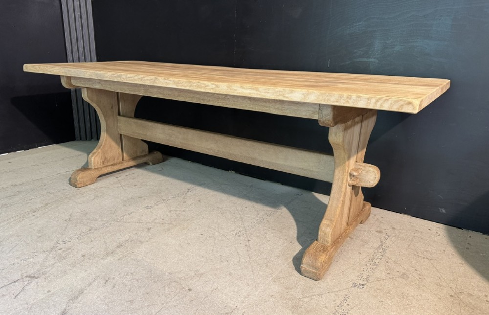 A long bleached oak dining table featuring a solid rectangular top supported by curved trestle legs connected by a central stretcher beam. View II