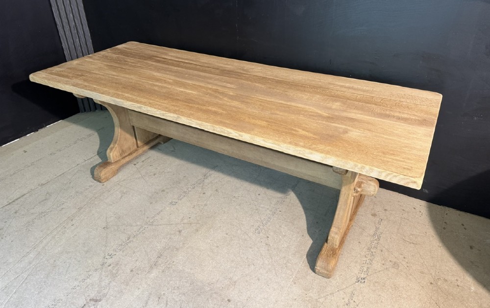 A long bleached oak dining table featuring a solid rectangular top supported by curved trestle legs connected by a central stretcher beam. View III