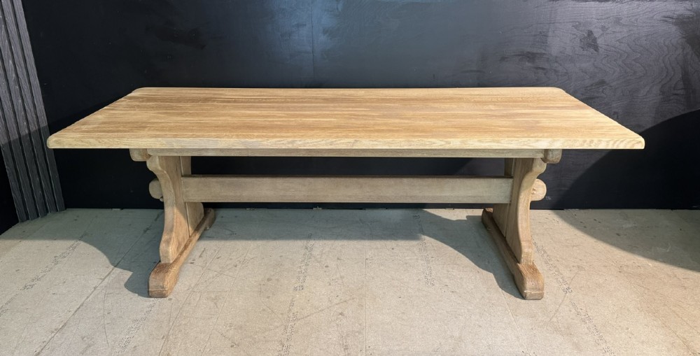 A long bleached oak dining table featuring a solid rectangular top supported by curved trestle legs connected by a central stretcher beam. View IV