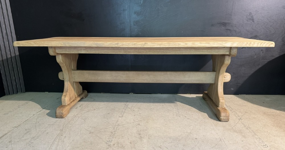 A long bleached oak dining table featuring a solid rectangular top supported by curved trestle legs connected by a central stretcher beam. View V