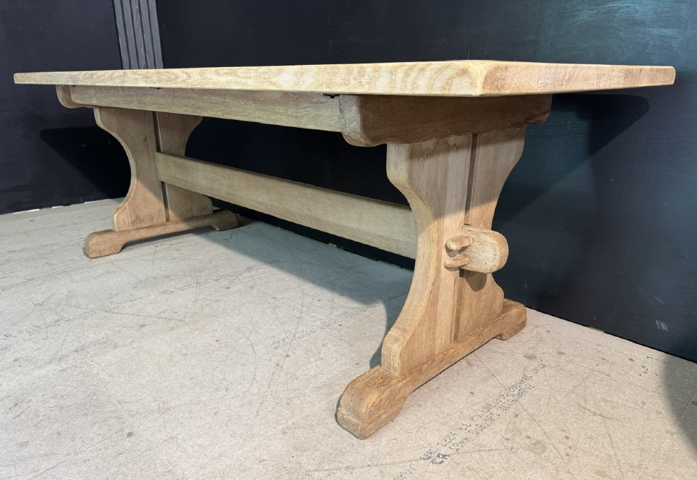 A long bleached oak dining table featuring a solid rectangular top supported by curved trestle legs connected by a central stretcher beam. View VI