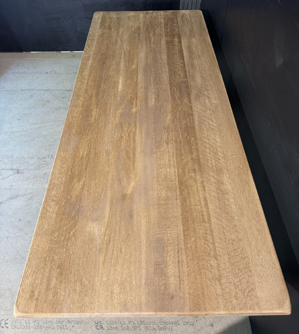 A long bleached oak dining table featuring a solid rectangular top supported by curved trestle legs connected by a central stretcher beam. View VII