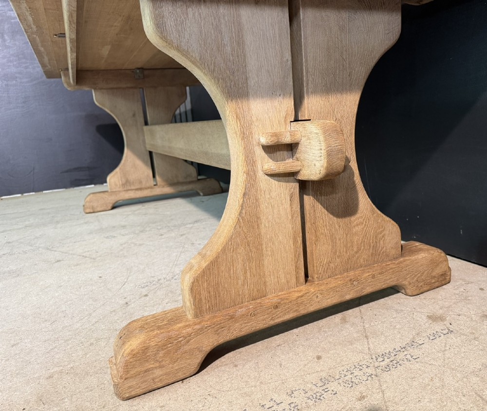 A long bleached oak dining table featuring a solid rectangular top supported by curved trestle legs connected by a central stretcher beam. View IX