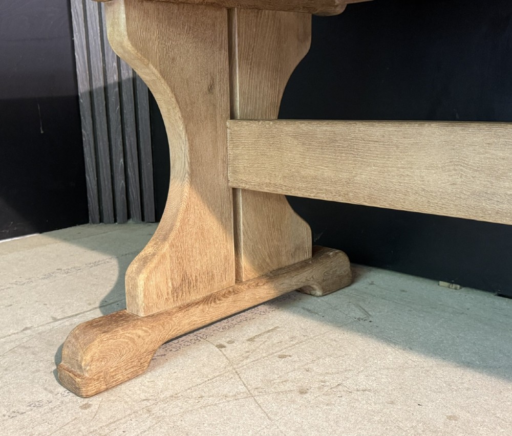 A long bleached oak dining table featuring a solid rectangular top supported by curved trestle legs connected by a central stretcher beam. View XI
