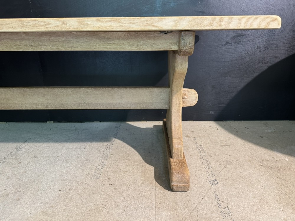 A long bleached oak dining table featuring a solid rectangular top supported by curved trestle legs connected by a central stretcher beam. View XII