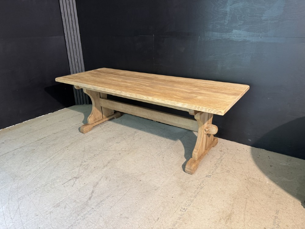 A long bleached oak dining table featuring a solid rectangular top supported by curved trestle legs connected by a central stretcher beam. View XV