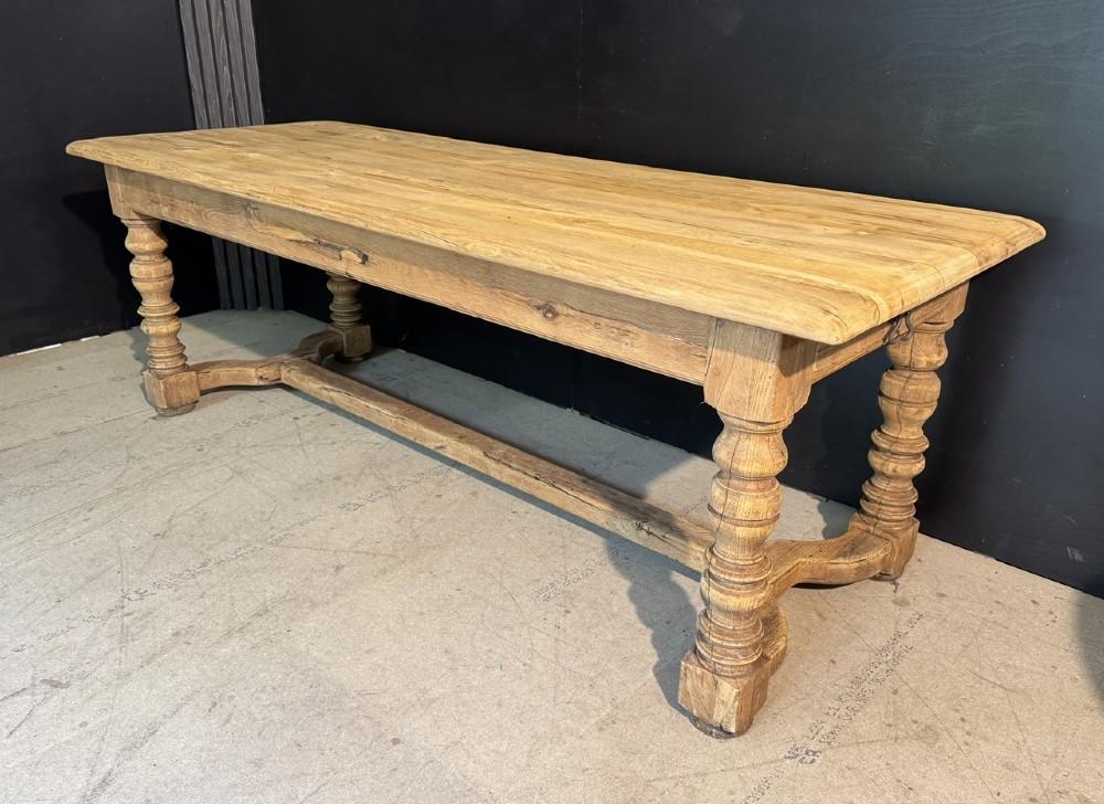 A long light oak French farmhouse table featuring a thick plank top, two frieze drawers, thick turned legs, and an H-stretcher base.