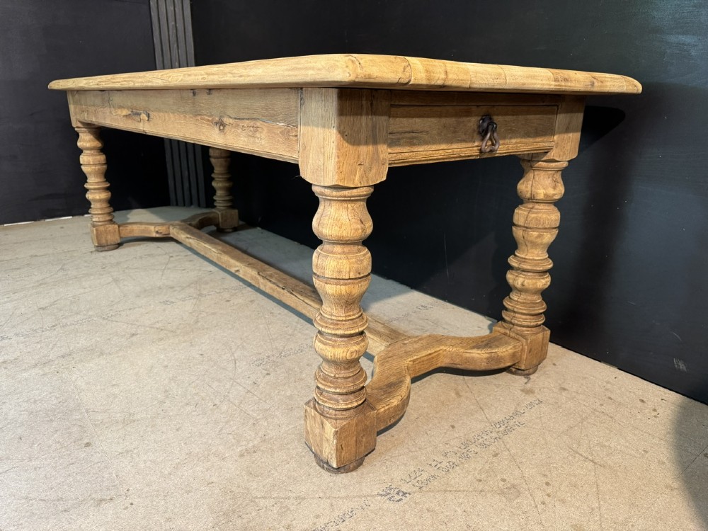 A long light oak French farmhouse table featuring a thick plank top, two frieze drawers, thick turned legs, and an H-stretcher base. View X