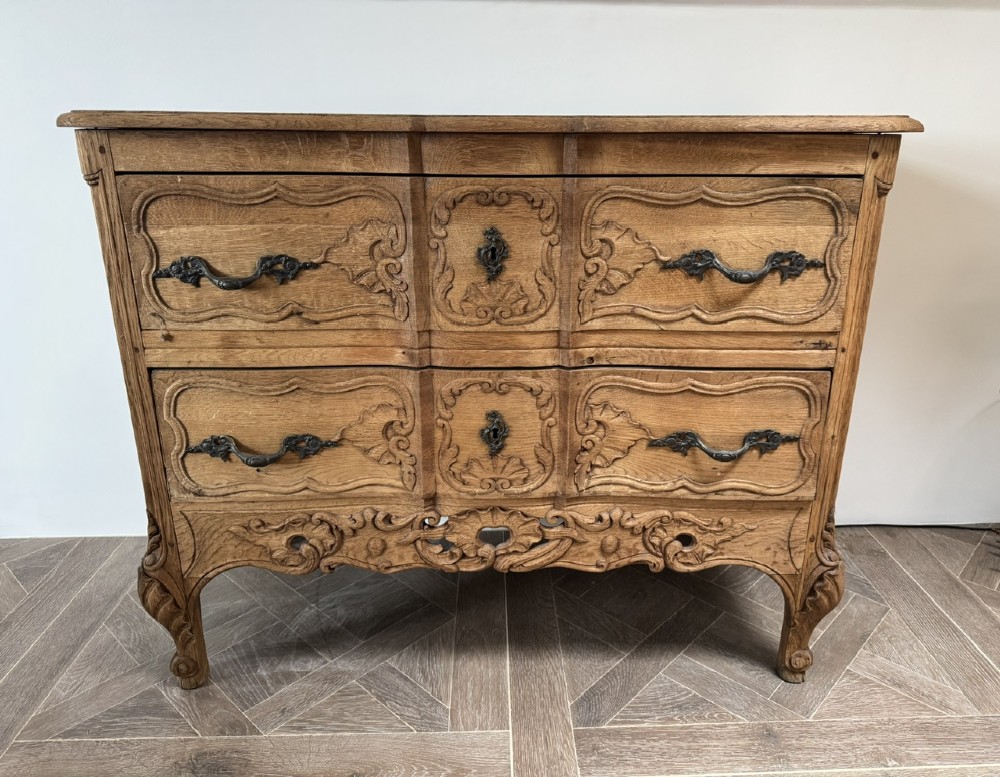 A French bleached oak two-drawer commode featuring carved foliate panels, cabriole legs, and dark metal handles.