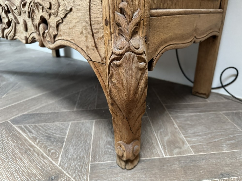 A French bleached oak two-drawer commode featuring carved foliate panels, cabriole legs, and dark metal handles. View VII