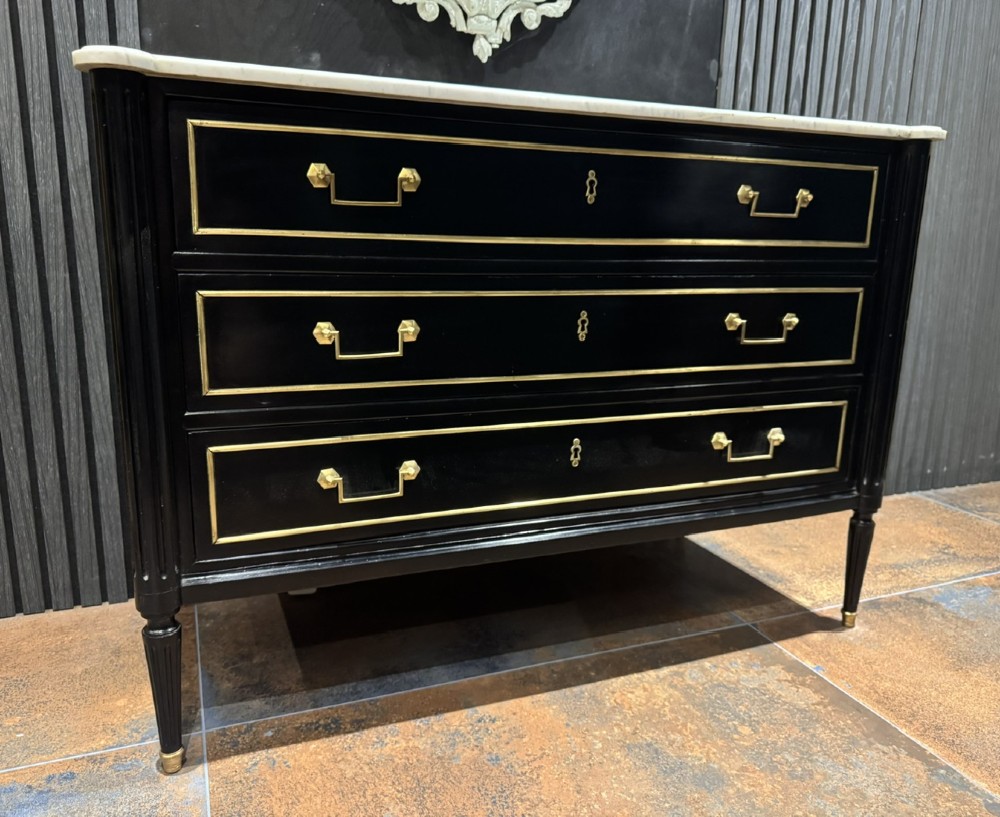 An ebonised Louis XVI style chest of drawers featuring three long drawers with brass rectangular mouldings, geometric handles, fluted tapered legs, and a white marble top. View III