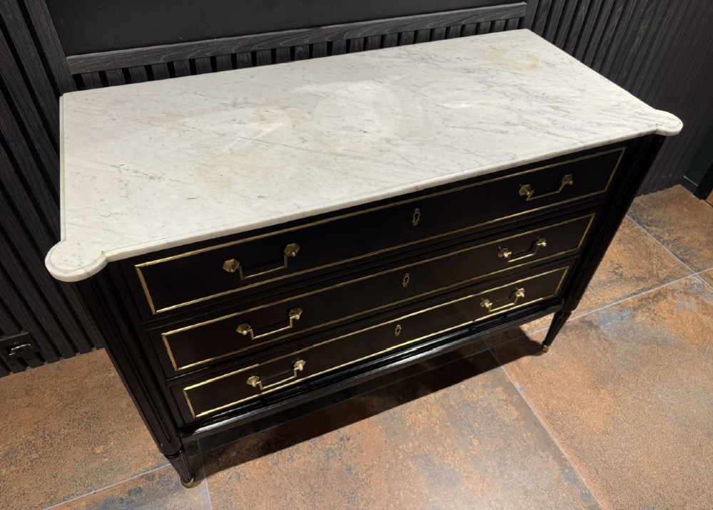 An ebonised Louis XVI style chest of drawers featuring three long drawers with brass rectangular mouldings, geometric handles, fluted tapered legs, and a white marble top. View IV