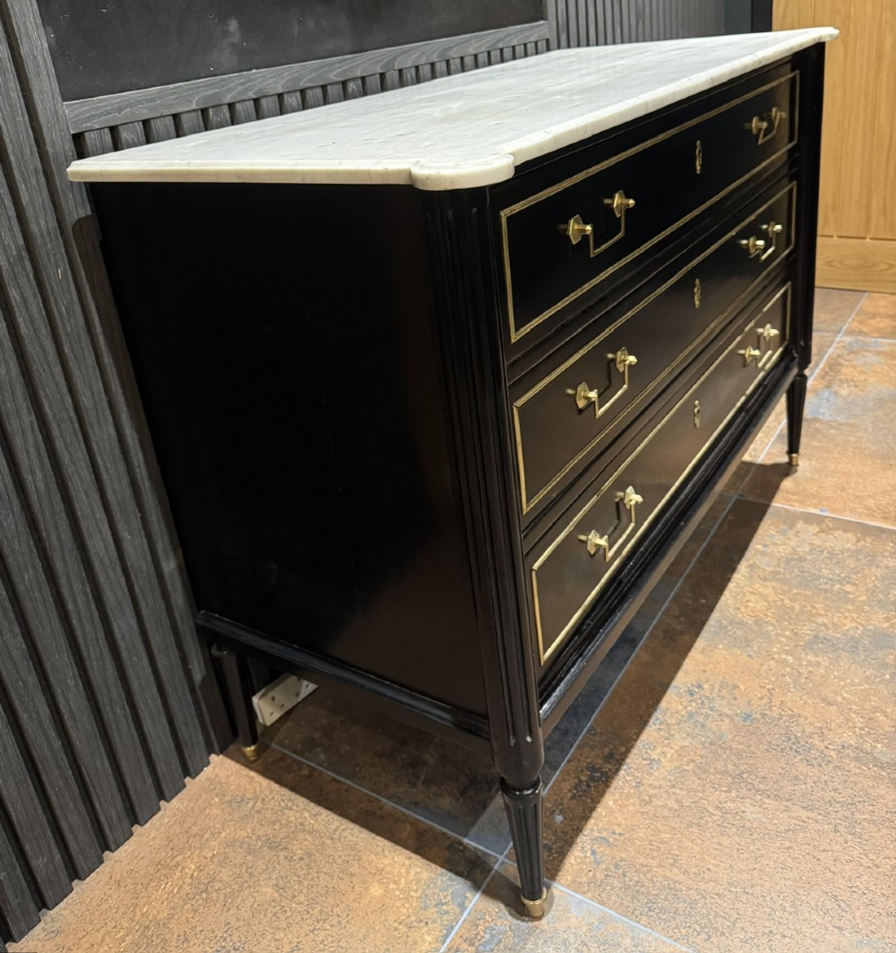 An ebonised Louis XVI style chest of drawers featuring three long drawers with brass rectangular mouldings, geometric handles, fluted tapered legs, and a white marble top. View V